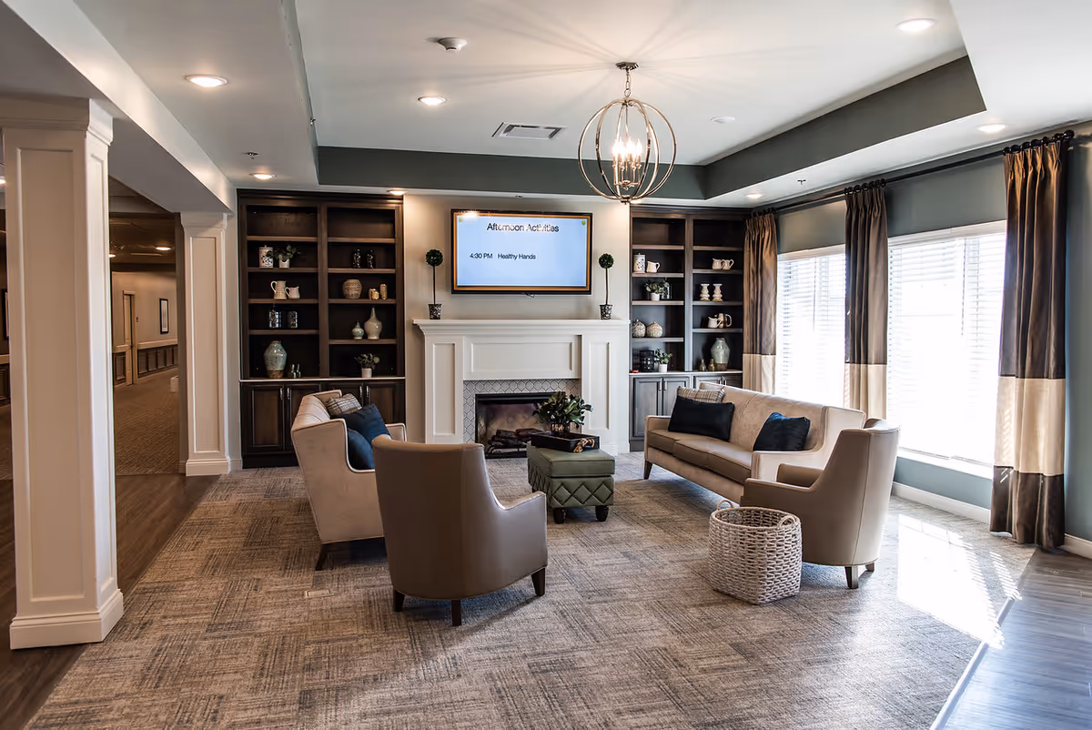 Bright senior living lounge with beige sofas, chairs, fireplace, shelves, and large windows.