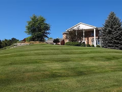Brick building with columns perched on a grassy hill, surrounded by trees beneath a clear blue sky.