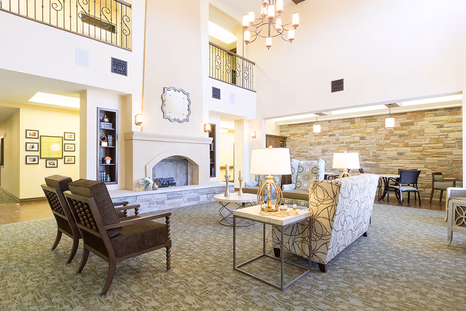 A spacious and well-lit senior living facility common area with a high ceiling, featuring a fireplace with a decorative mirror above it, comfortable seating including patterned armchairs and wooden chairs, side tables with lamps, and a stone accent wall in the background with additional seating and hanging lights.