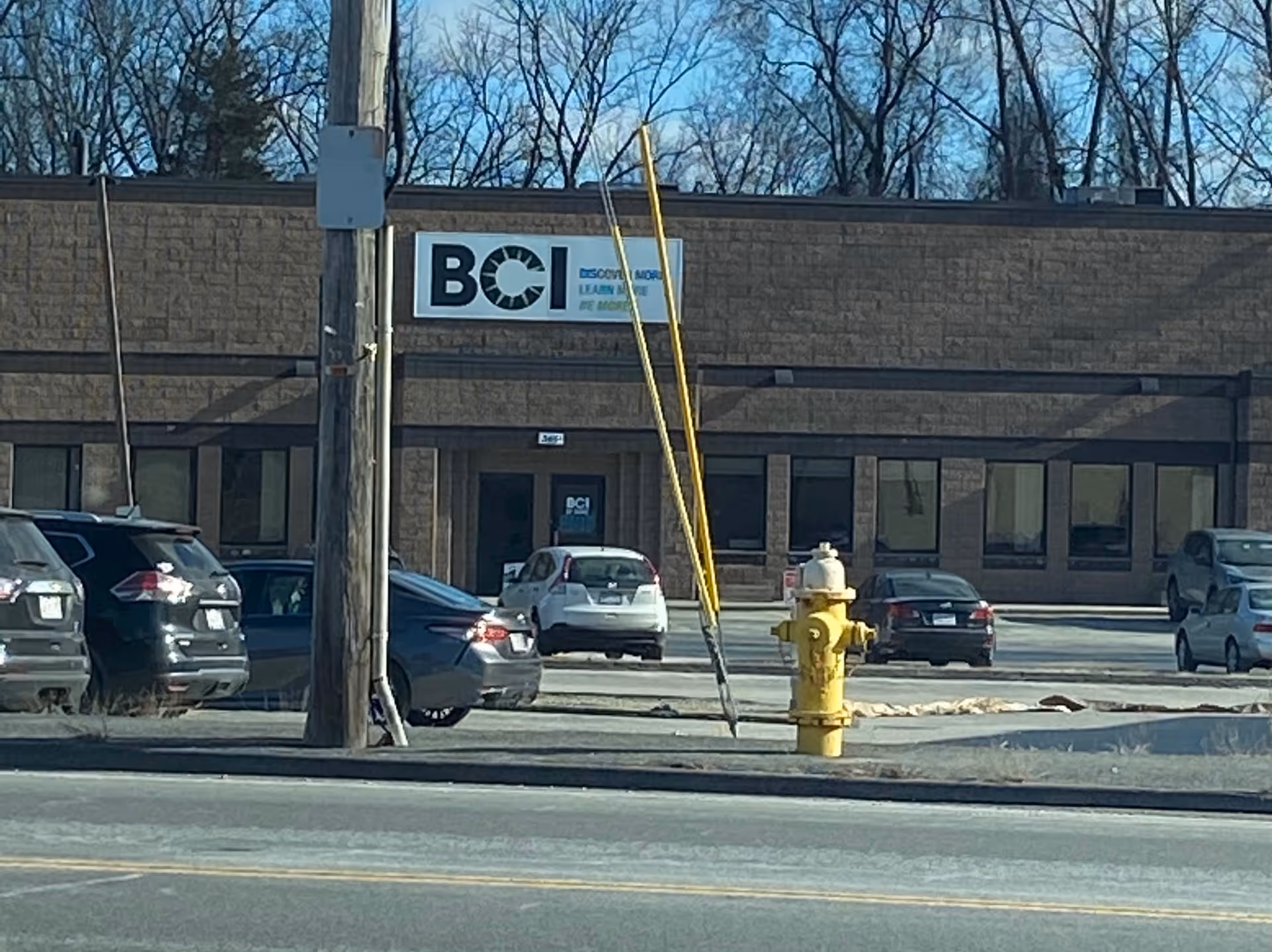 Exterior of a single-story building with a BCI sign, parked cars, and a yellow fire hydrant.