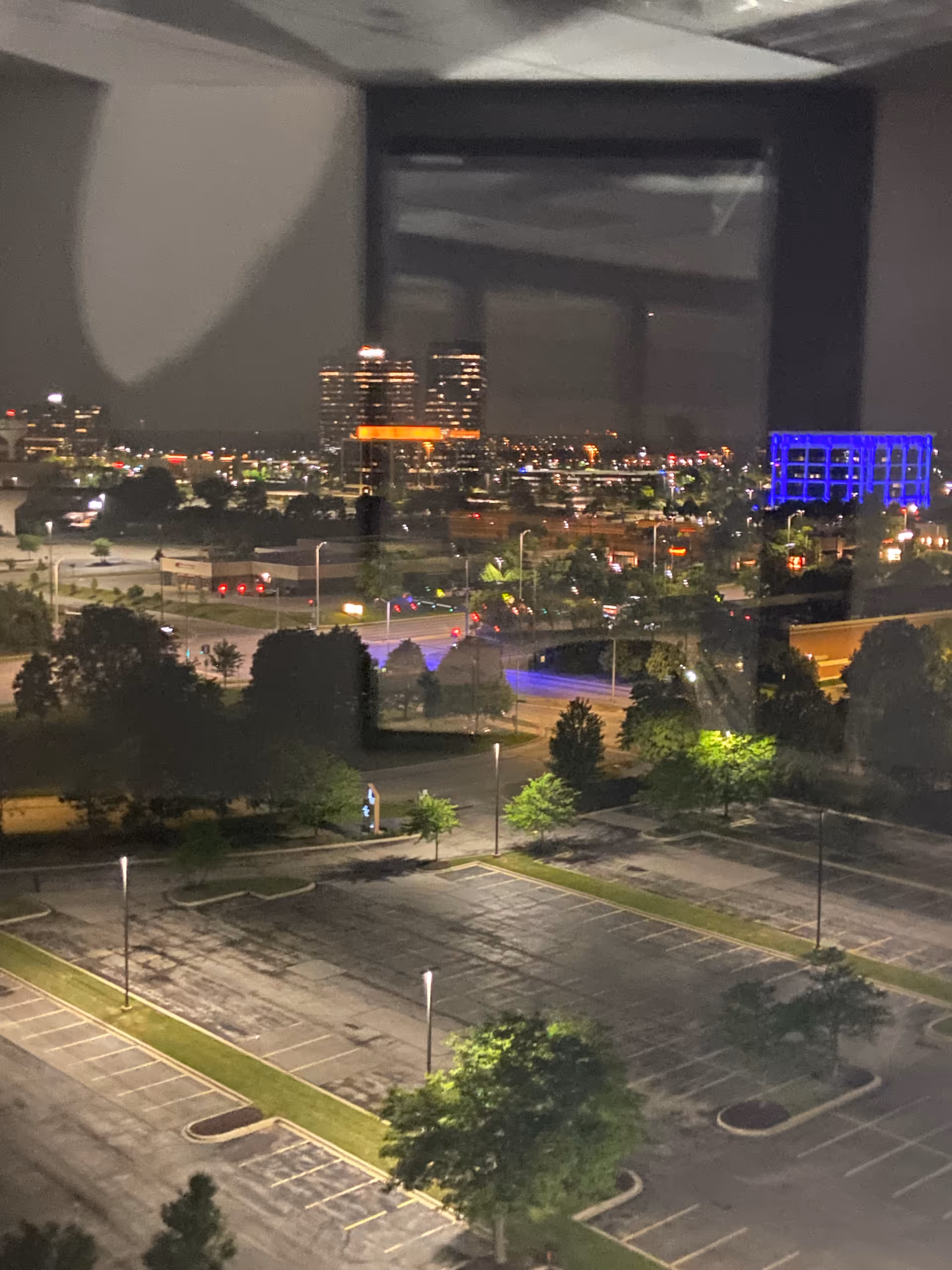Nighttime cityscape viewed from inside through a window, showing a parking lot and lit buildings.