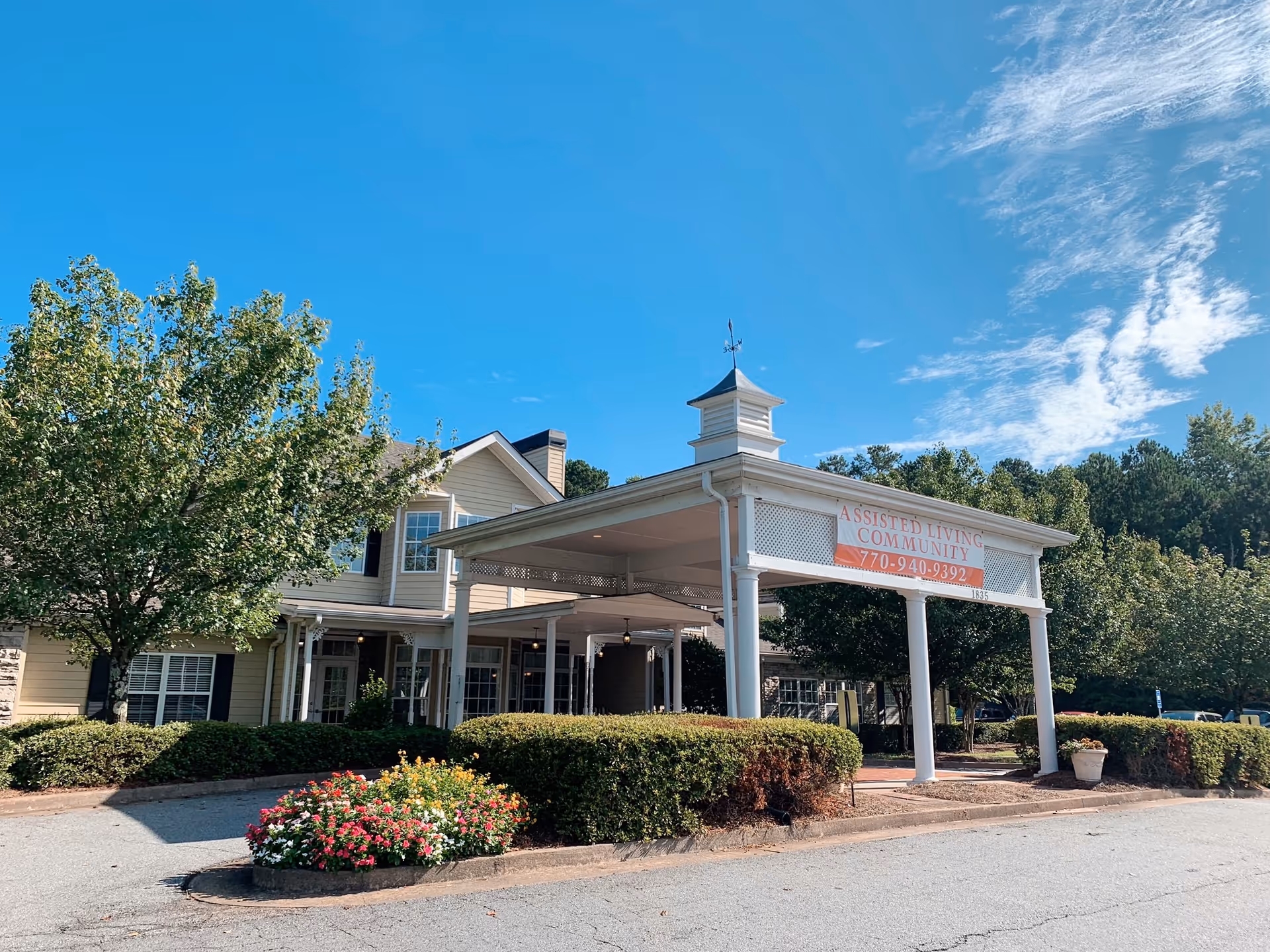 Exterior view of Sunabella of Towne Lake assisted living community building with a covered entrance, surrounded by trees, bushes, and flowers under a clear blue sky.