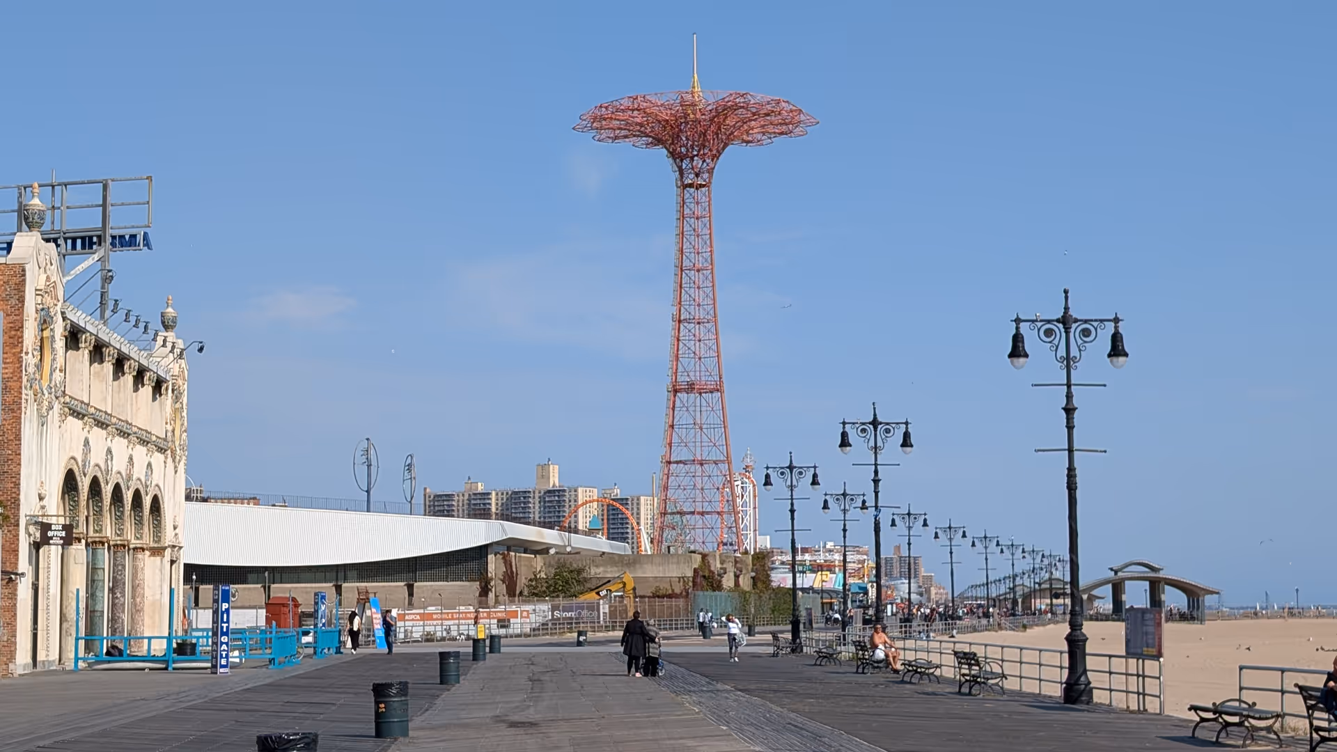 Seaside boardwalk with benches, ornate lampposts, and a red tower along the beach.