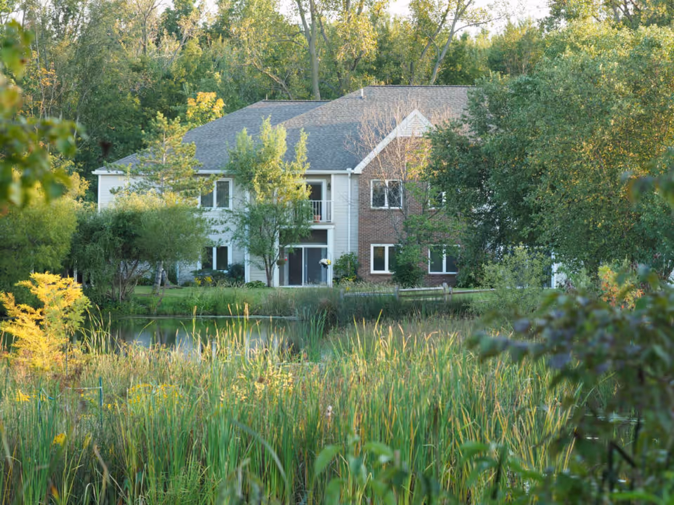 A two-story residential building partially obscured by trees and greenery, with a small pond and tall grasses in the foreground, surrounded by lush vegetation and trees.