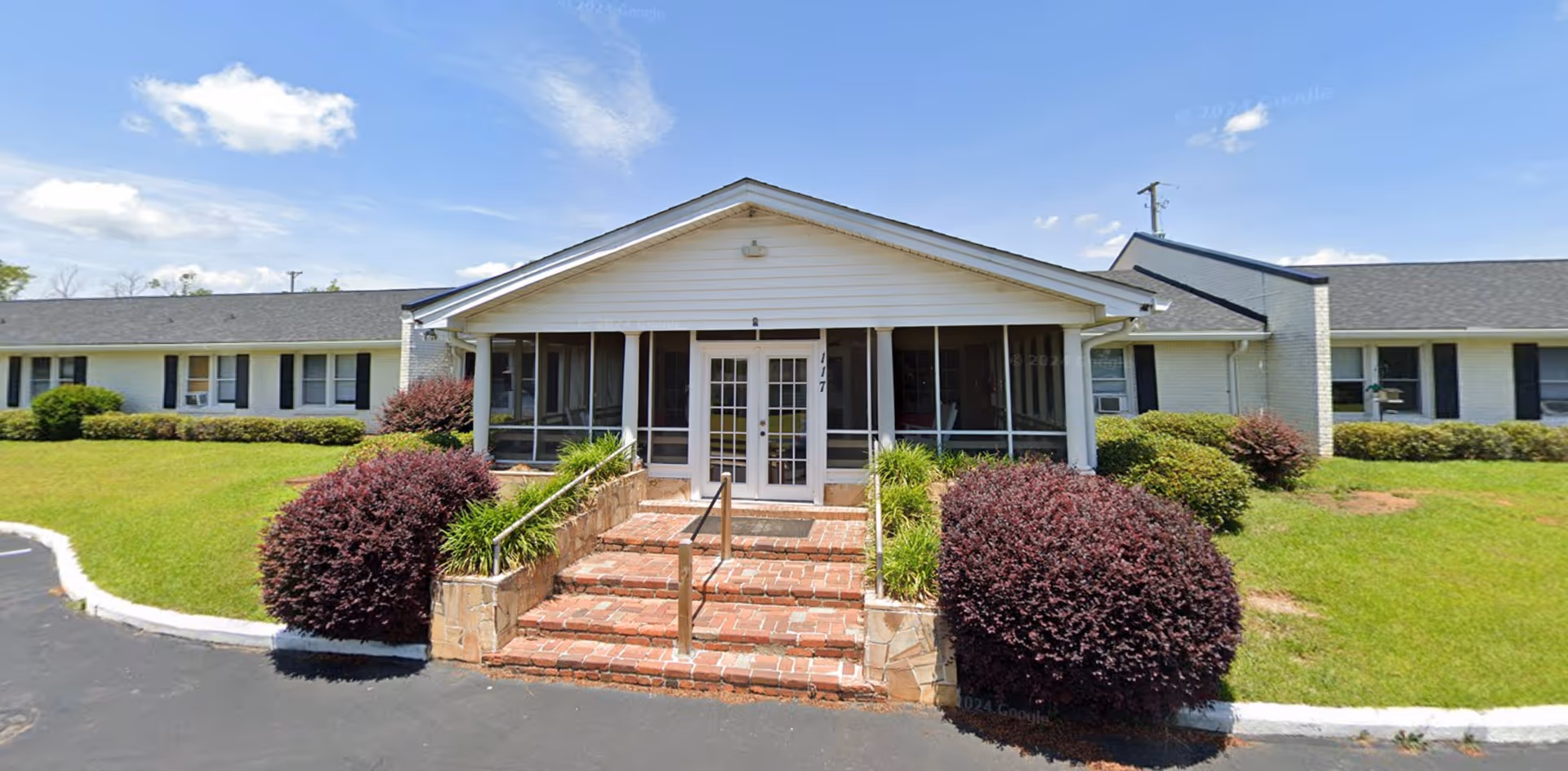 Front exterior view of Ridgeway Manor Healthcare Center showing a single-story building with white siding, black shutters, and a central entrance with steps and handrails. The entrance has a covered porch with screened windows. There are well-maintained bushes and green grass surrounding the building under a blue sky with some clouds.