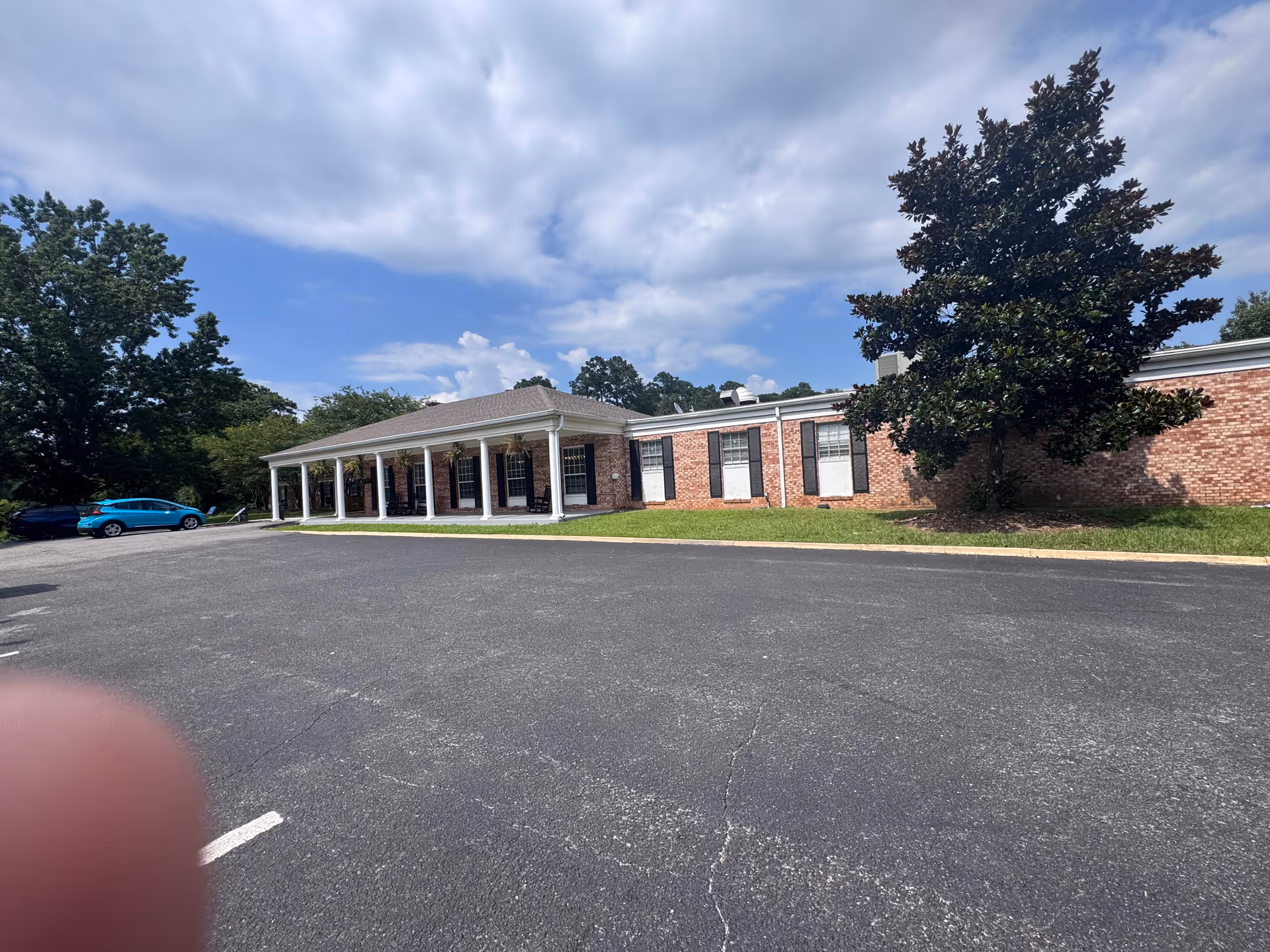 Brick building with a white-columned porch entrance, adjacent parking lot and trees.