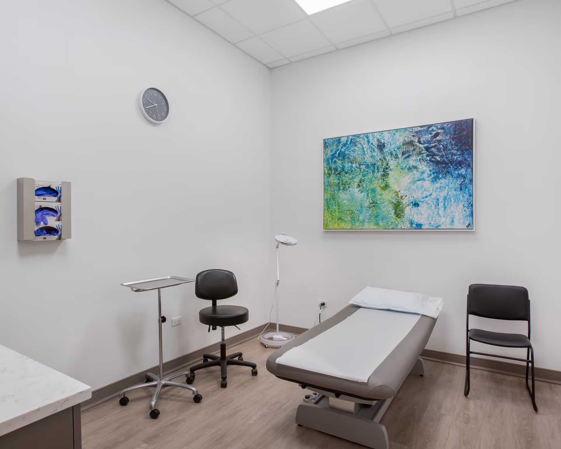 Examination room featuring a patient bed, rolling stool, chair, lamp, and abstract wall art.