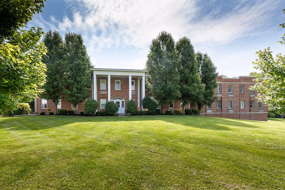 Front exterior view of a two-story brick building with white columns at the entrance, surrounded by green trees and a well-maintained lawn under a partly cloudy sky.