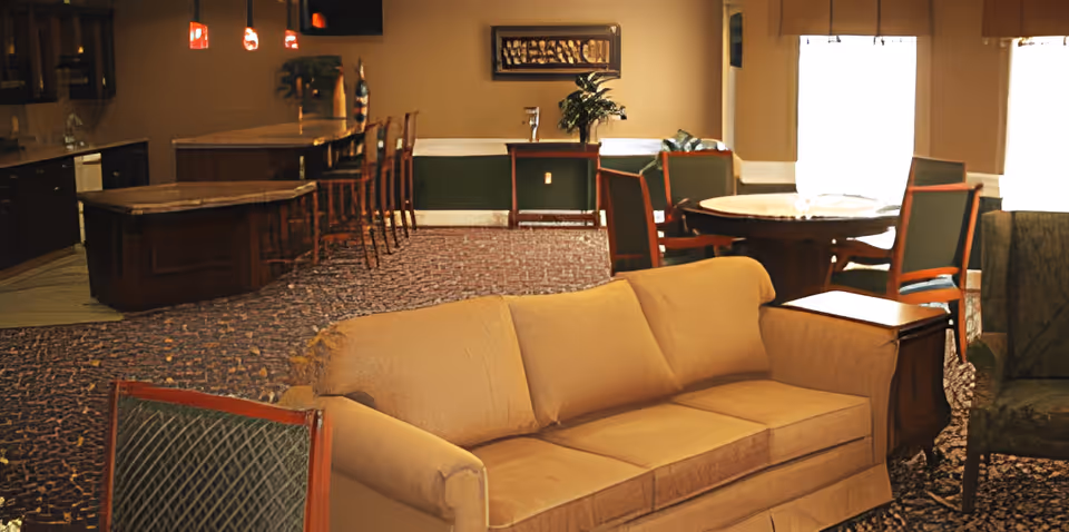 A communal lounge with a beige sofa, upholstered chairs, a round dining table, and a bar area in the background.