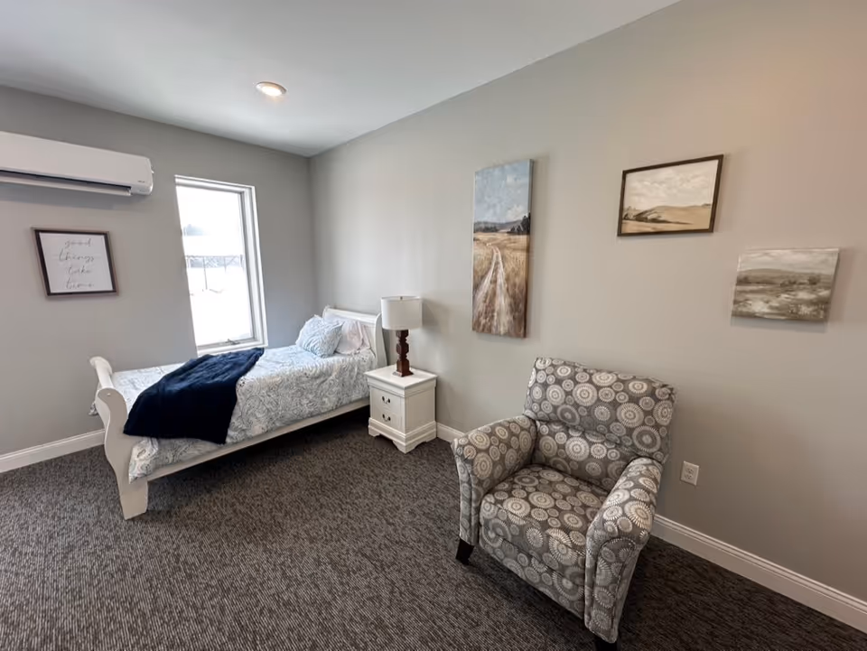 Cozy bedroom in a senior living facility with a bed, chair, and wall art.