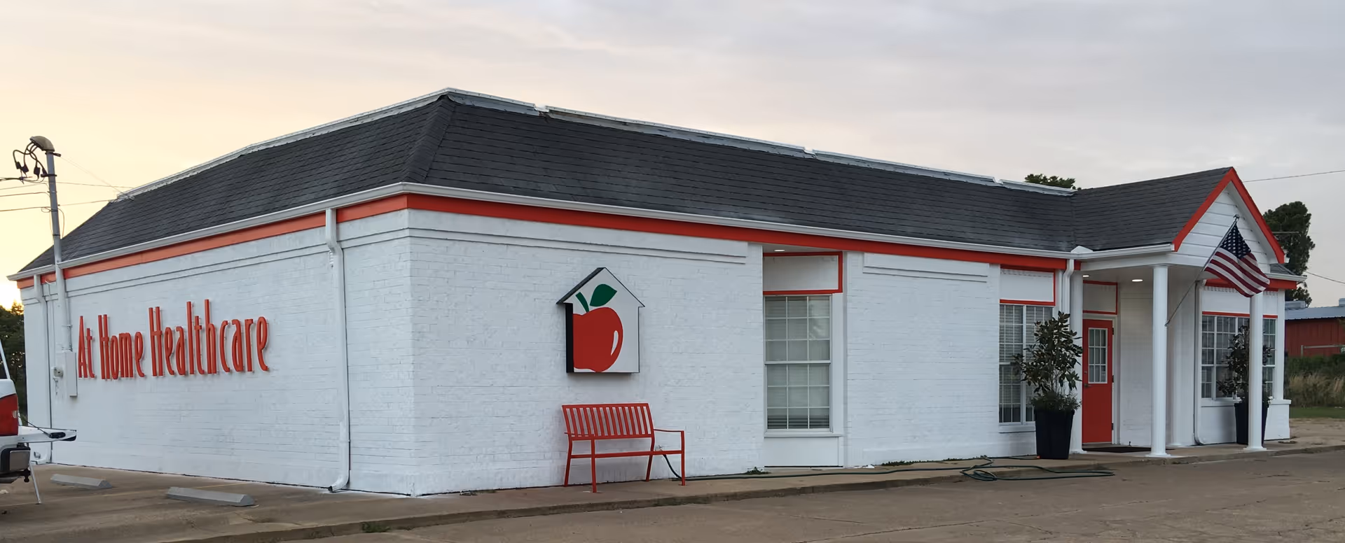 Exterior of a white building labeled At Home Healthcare with red trim and bench