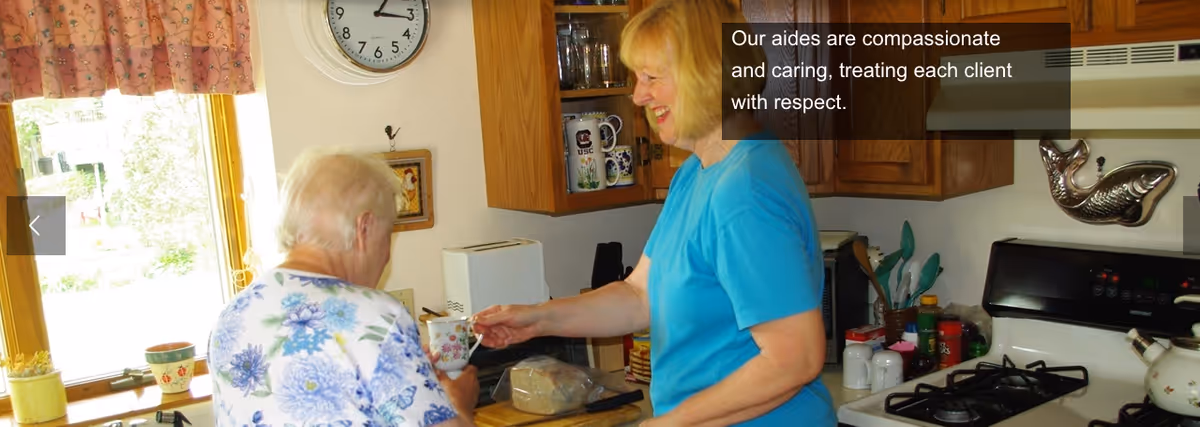 Caregiver hands a mug to an elderly woman in a bright kitchen.