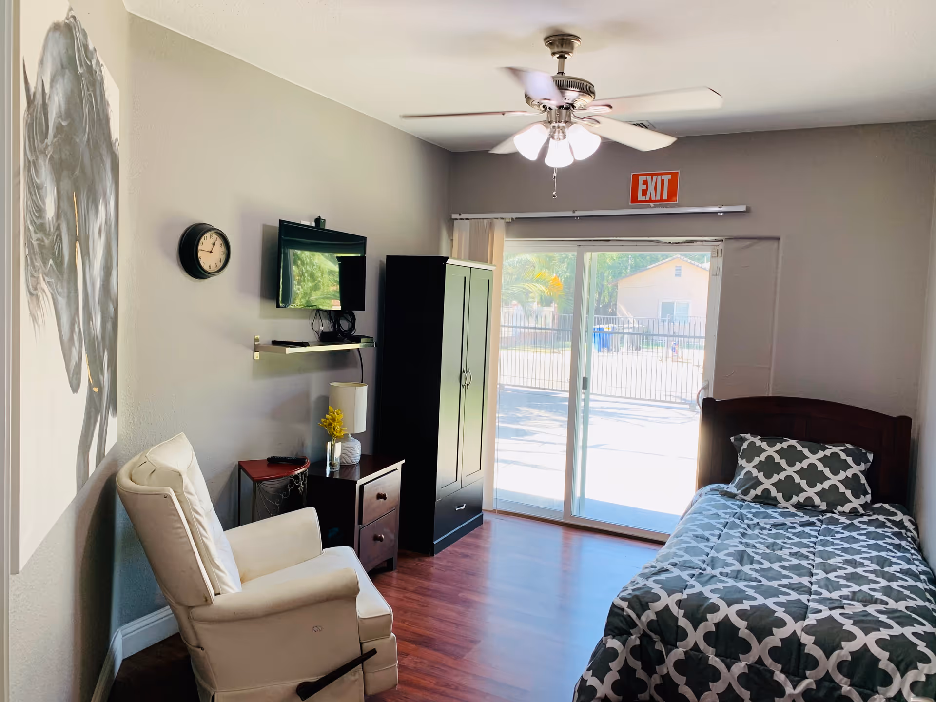 A furnished senior living bedroom with bed, recliner, wardrobe, TV, and sliding glass door.