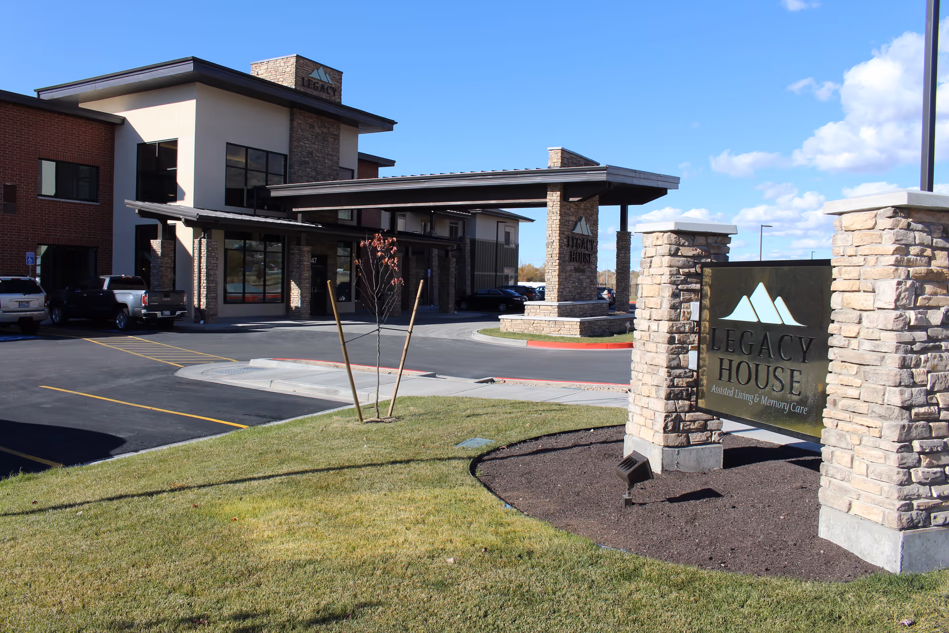 Exterior view of Legacy House of Park Lane, a modern assisted living and memory care facility with stone and brick architecture, a covered entrance, parking spaces, and a sign displaying the facility's name.