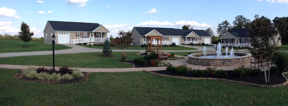 Single-story cottages around a landscaped courtyard with a central stone fountain, walkways, and benches under a blue sky.