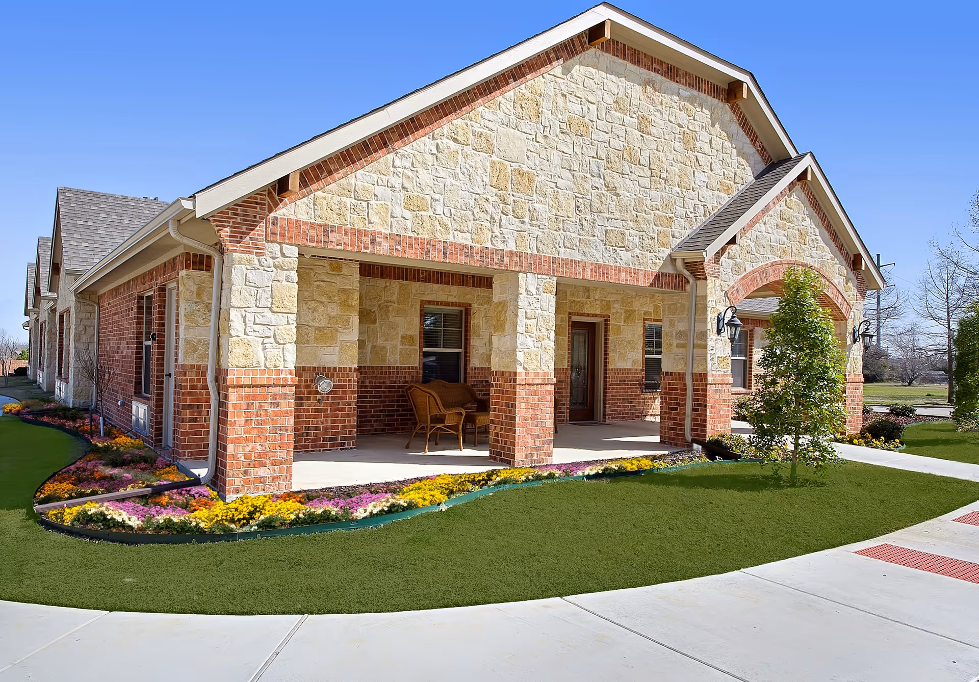 Stone and brick senior living facility front entrance with landscaped flower beds and walkway.