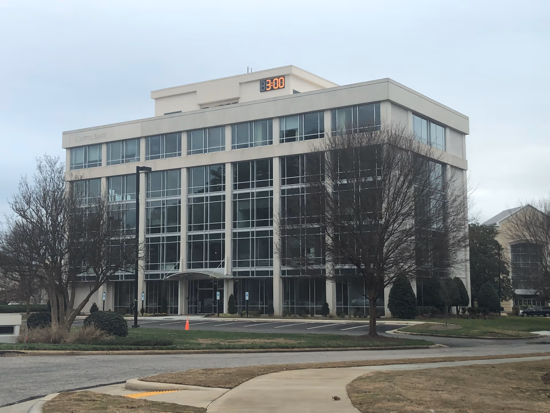 Modern glass-front building with leafless trees and a parking lot in front.