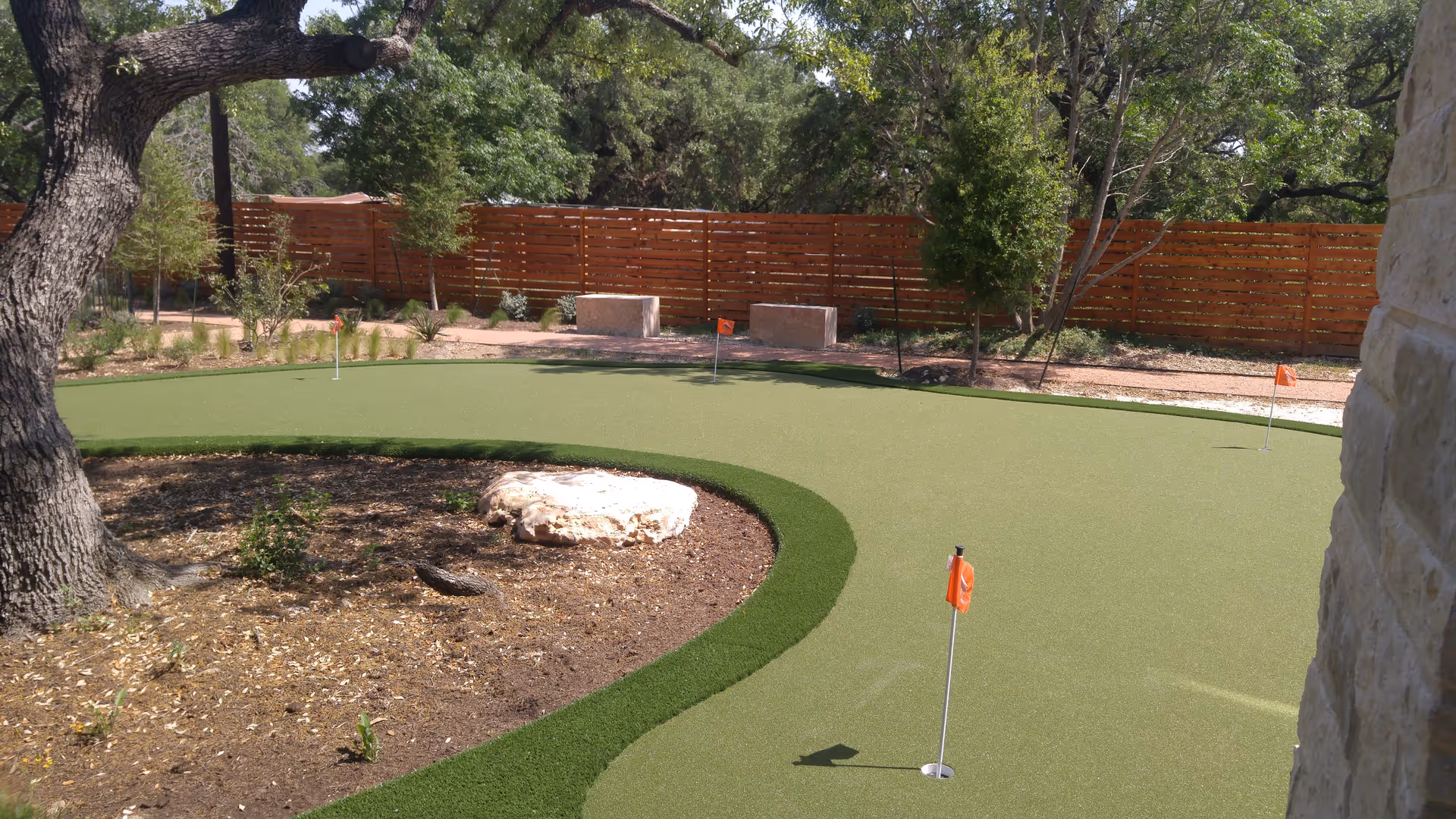 Outdoor putting green with several hole flags, surrounded by trees, a wooden fence and stone features.