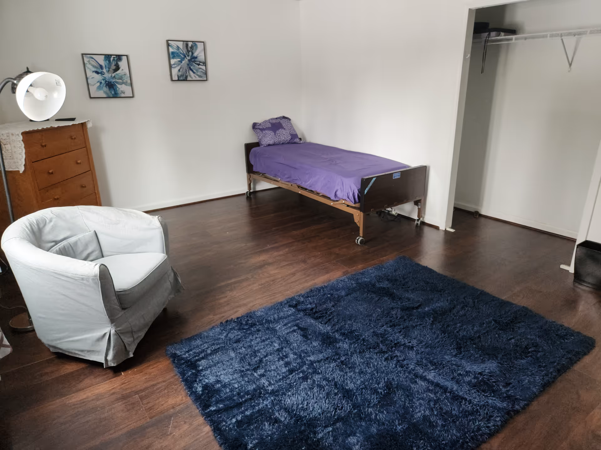 Bedroom with a single bed, purple linens, dresser, gray chair, and dark blue rug.