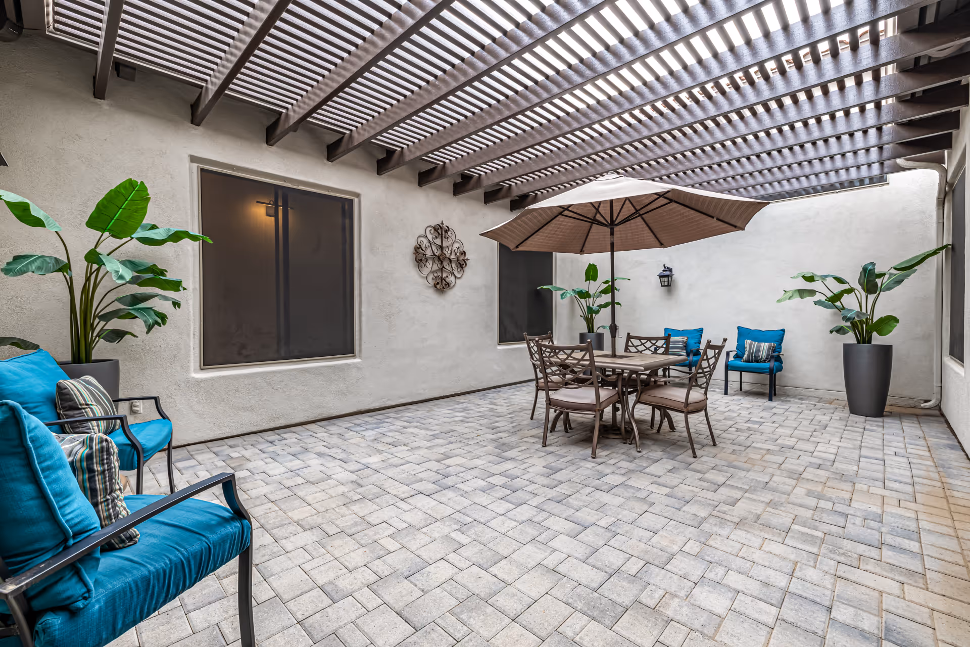 A spacious outdoor patio area with a pergola overhead providing partial shade. The patio is paved with light-colored bricks and features a round table with four chairs and a large umbrella. There are two blue cushioned chairs with striped pillows on the left and one blue cushioned chair on the right. Large potted plants are placed around the patio, and a decorative wall piece and a lantern-style light fixture are mounted on the beige walls.