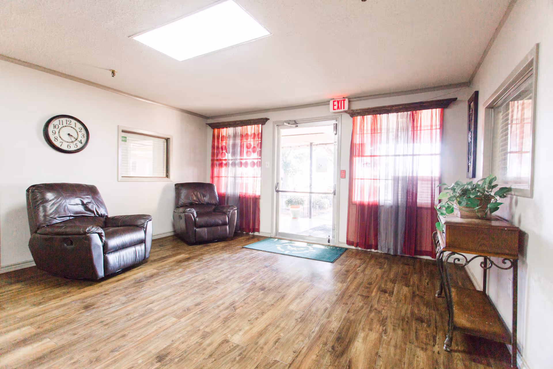 A spacious senior living lounge with two brown leather recliners, wooden floor, and red curtains.