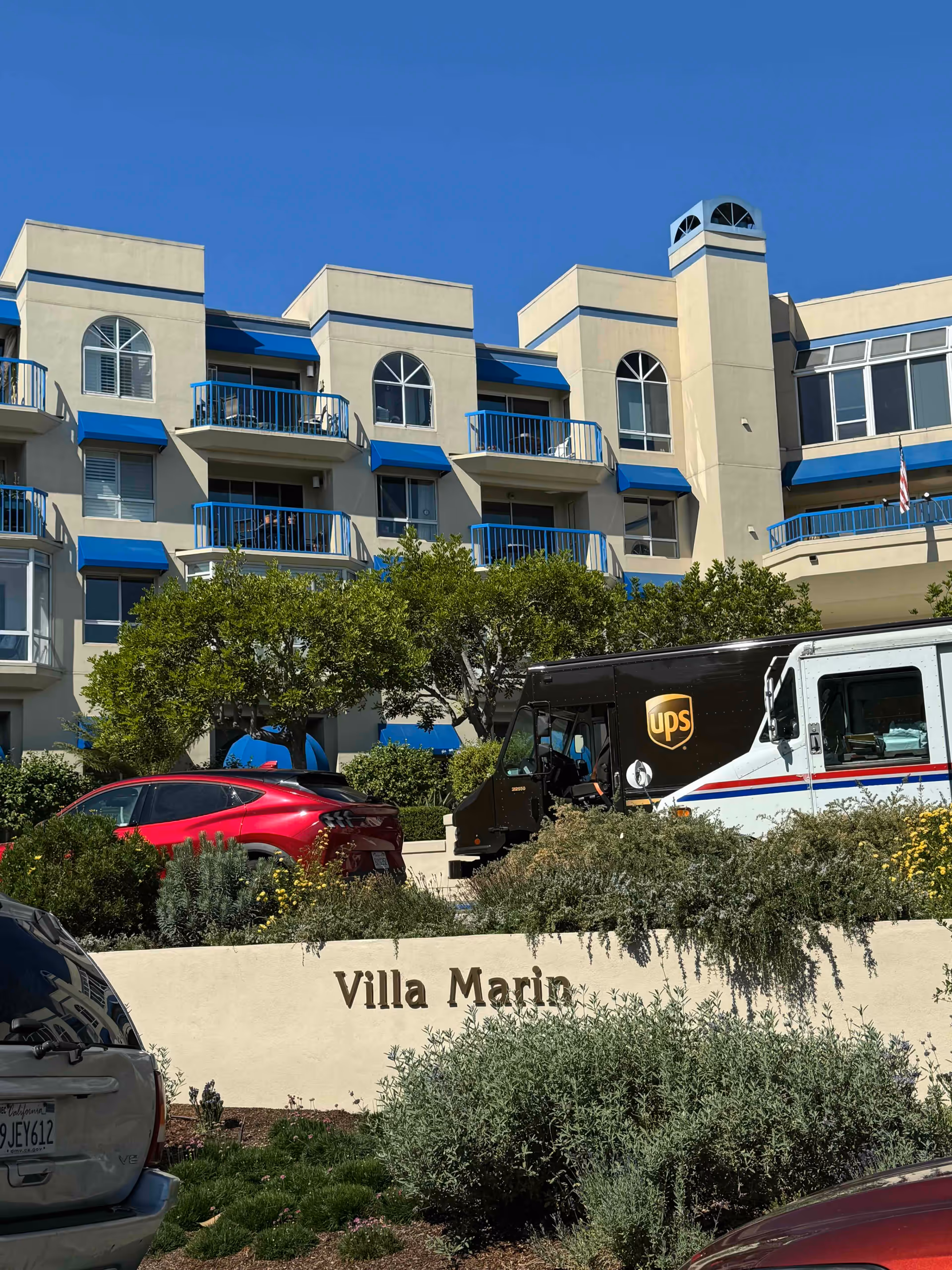 Front exterior of the Villa Marin building with blue-accented balconies, landscaping, and delivery vehicles parked in front.