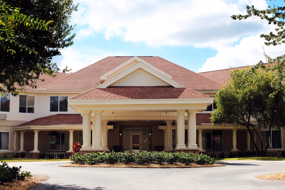 Front entrance of a two-story assisted living building with a covered portico, columns, and landscaped circular driveway.