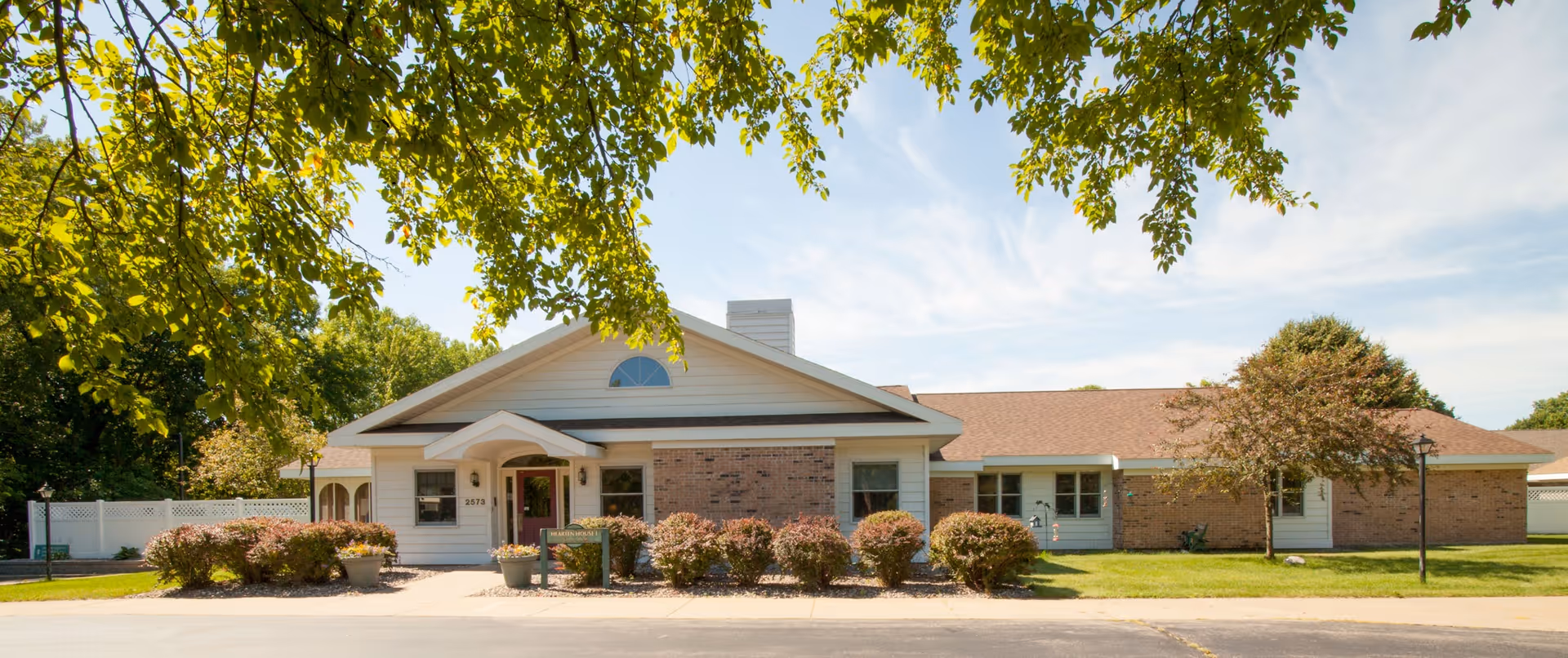 Single-story brick and siding building with a central entrance, landscaped shrubs and trees, and a clear blue sky.