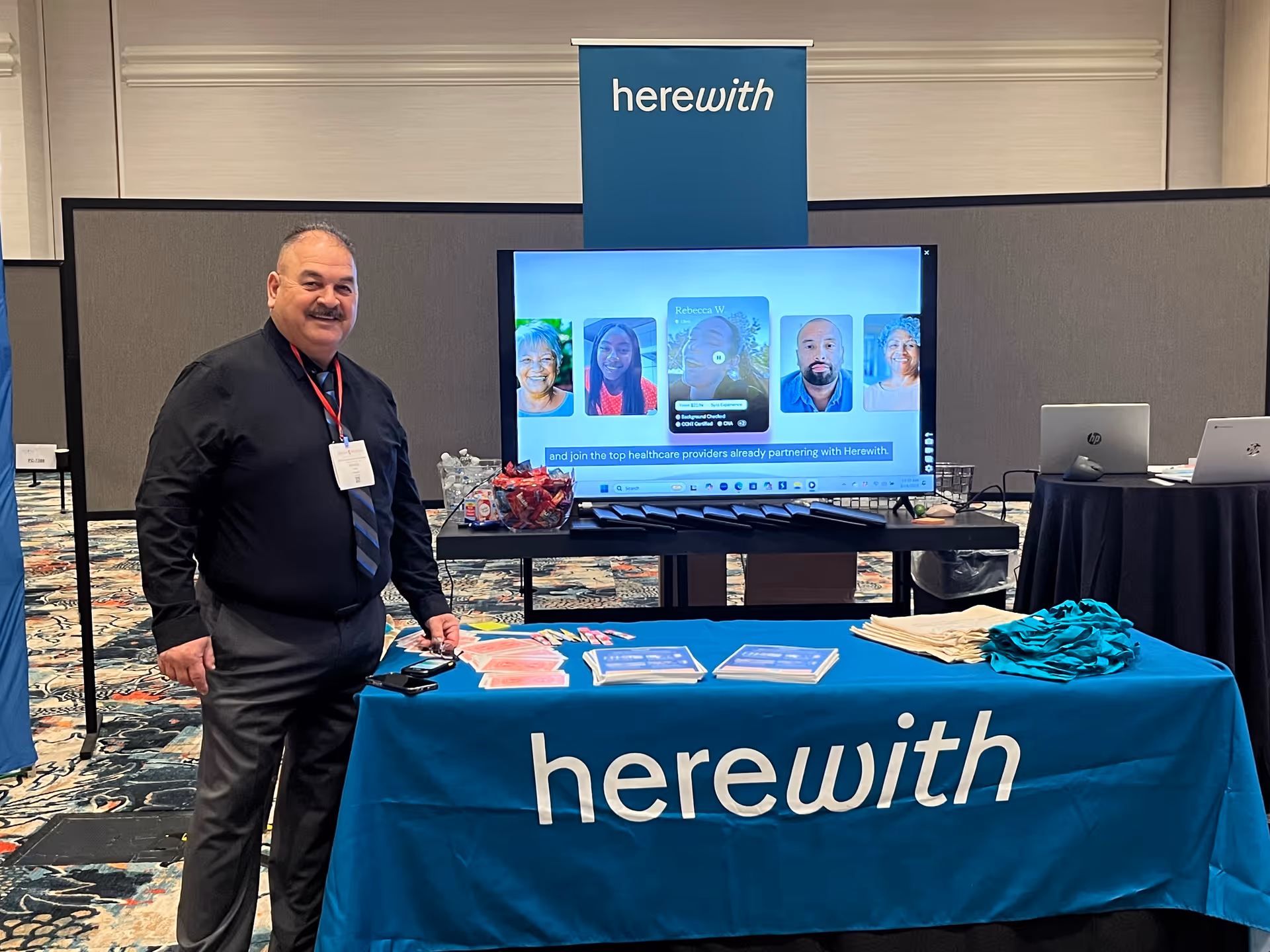 A man stands beside a blue Herewith booth with a large TV display.