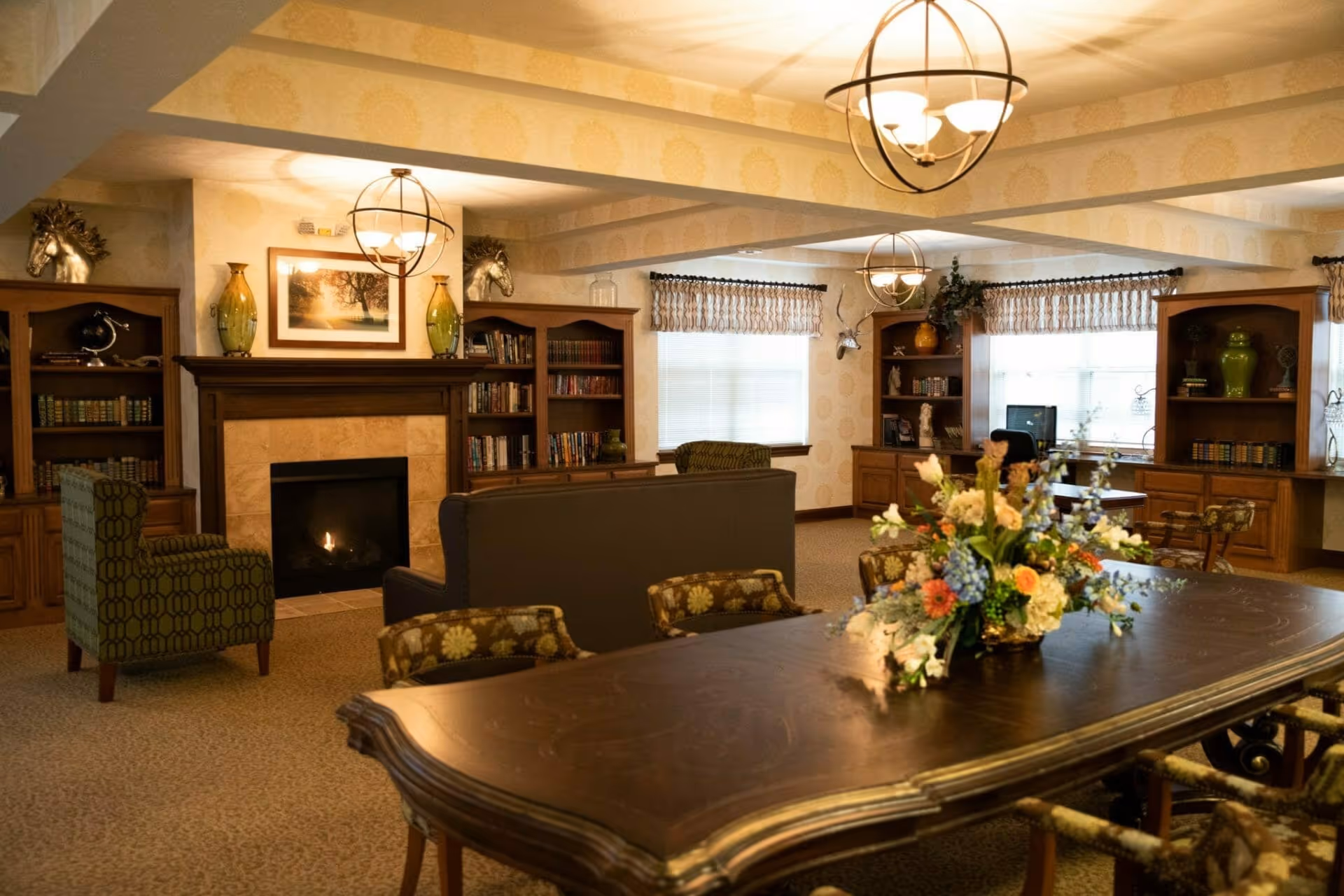 A warm common living room with a fireplace, bookshelves, seating, and a large table topped with a floral centerpiece.
