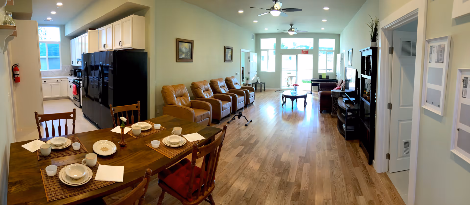 A spacious interior of an assisted living facility showing a dining area with a wooden table set for four, a kitchen with white cabinets and black appliances, and a living room with four brown recliner chairs, a coffee table, and a TV. The room has wooden flooring, light-colored walls, and large windows letting in natural light.