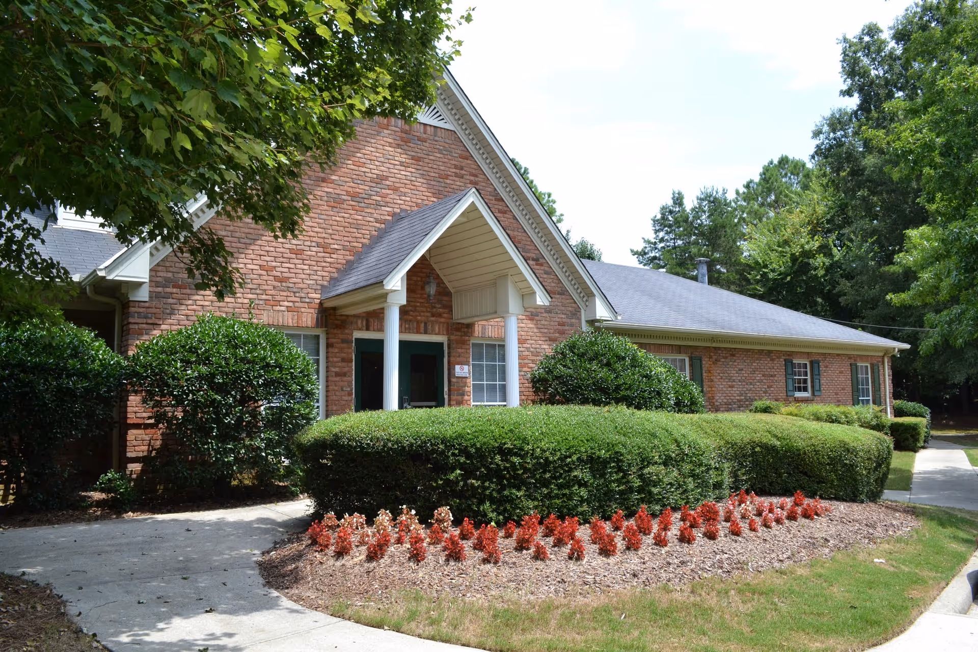 Exterior view of a single-story brick building with a gabled roof, surrounded by well-maintained bushes, red flowers, and trees. A concrete pathway leads to the entrance, which has a small covered porch supported by white columns.