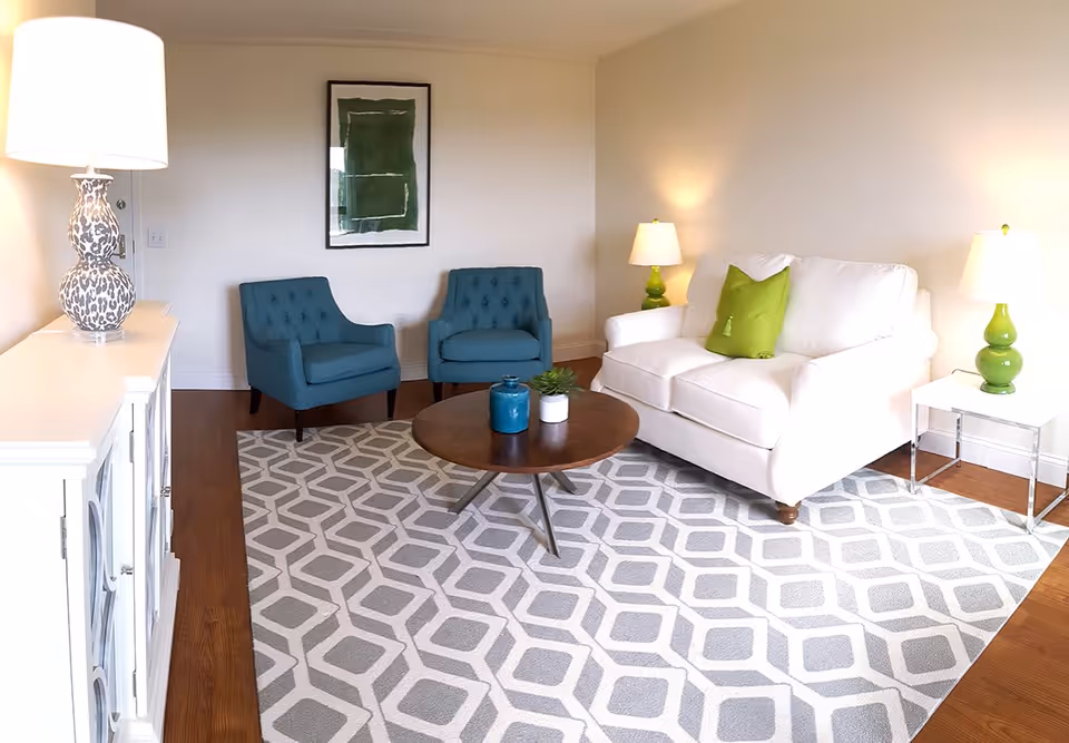 Bright living room with a white loveseat, two teal armchairs, a round wooden coffee table on a geometric rug, and green-accent lamps.