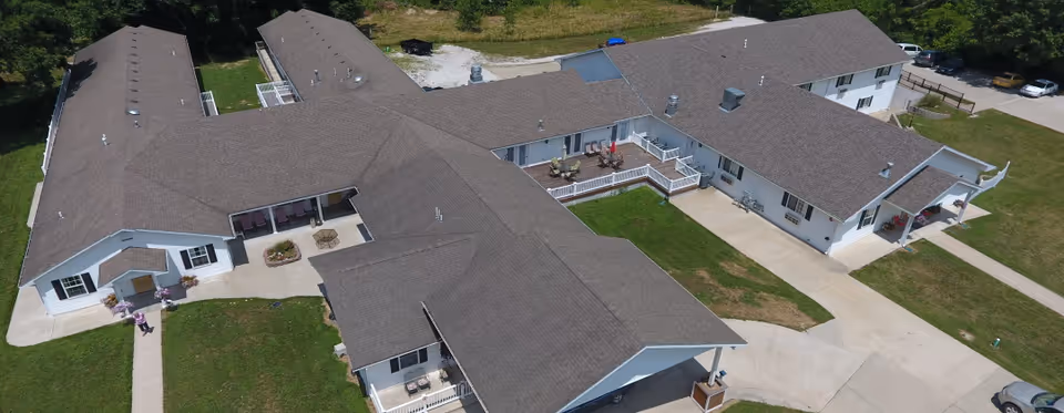 Aerial view of a single-story assisted living complex with interconnected pitched-roof wings, surrounding lawns, walkways, and a small courtyard patio.