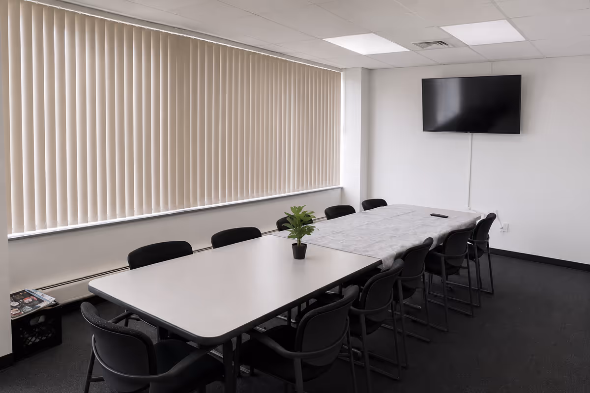 Rectangular conference table with chairs, blinds, and a wall-mounted TV in a bright meeting room.