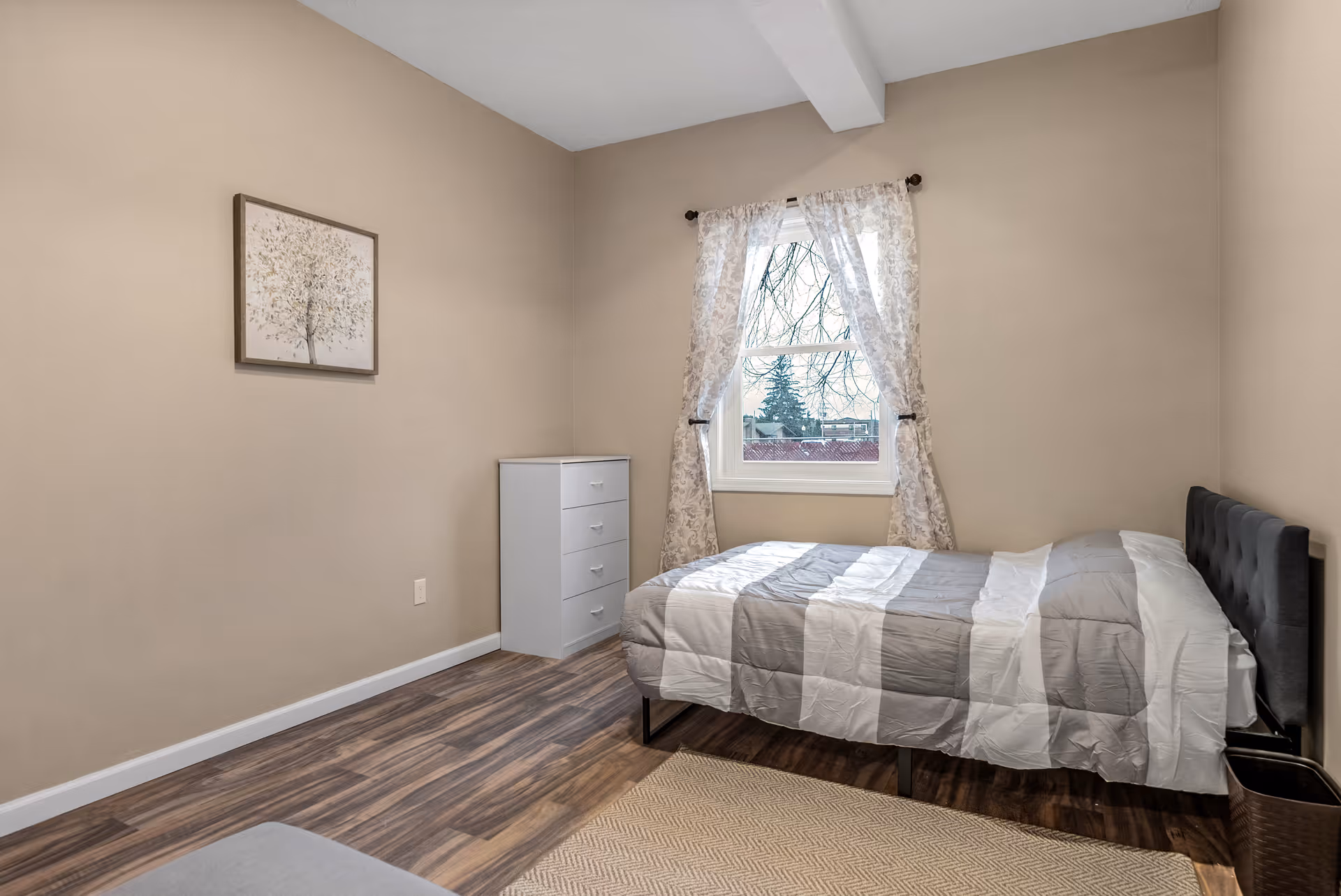 A simple bedroom with beige walls and wood flooring. The room features a single bed with a gray and white striped comforter, a white chest of drawers, a window with sheer patterned curtains, and a framed picture of a tree on the wall.