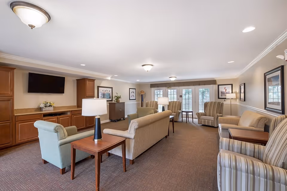 A spacious living room area in a senior living facility with multiple armchairs and sofas arranged around wooden side tables with lamps. The room features a wall-mounted flat-screen TV, wooden cabinetry, framed artwork, and large windows with glass doors letting in natural light.