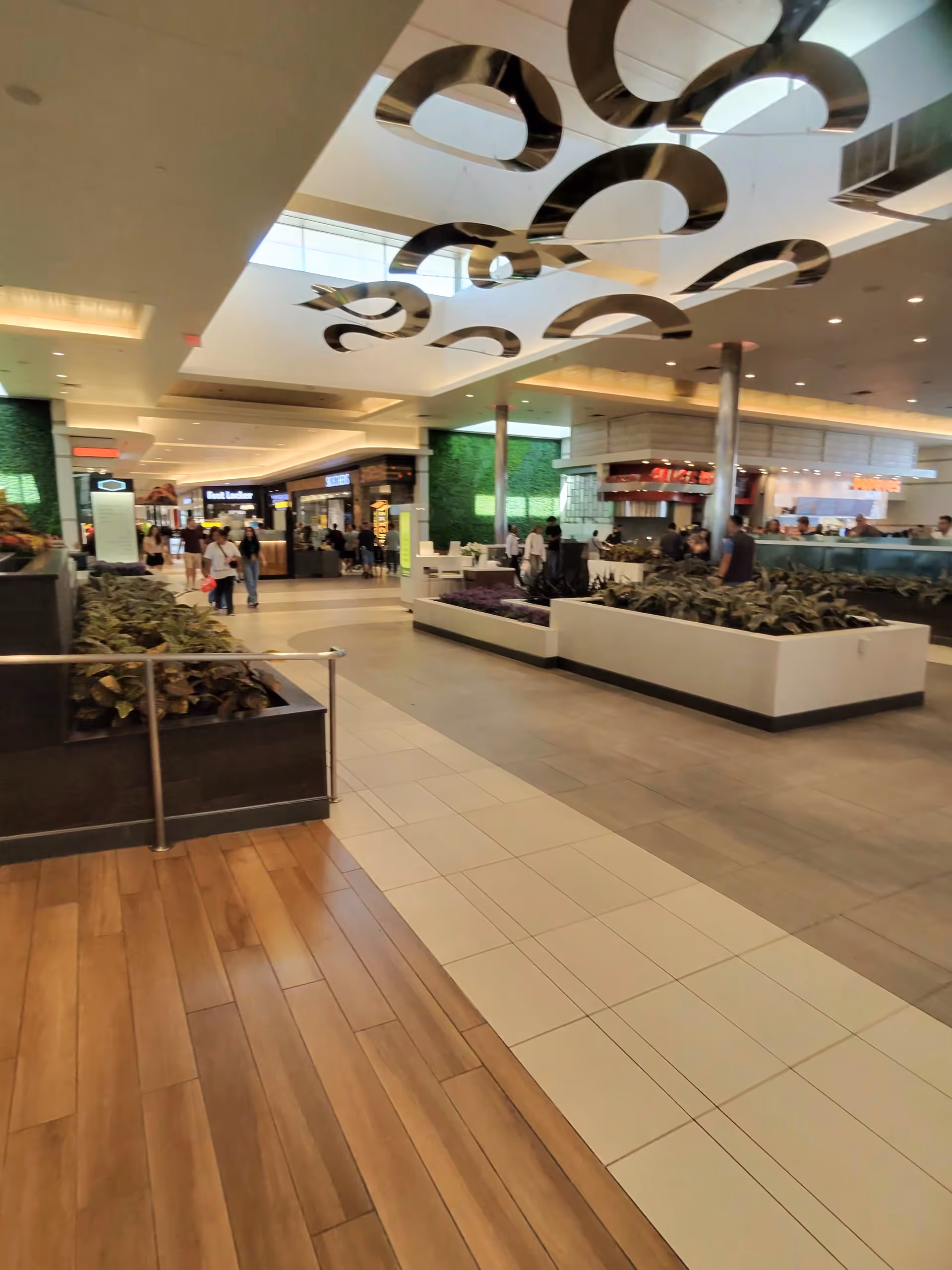 Indoor mall corridor with planters, storefronts, and people walking beneath a decorative ceiling sculpture.