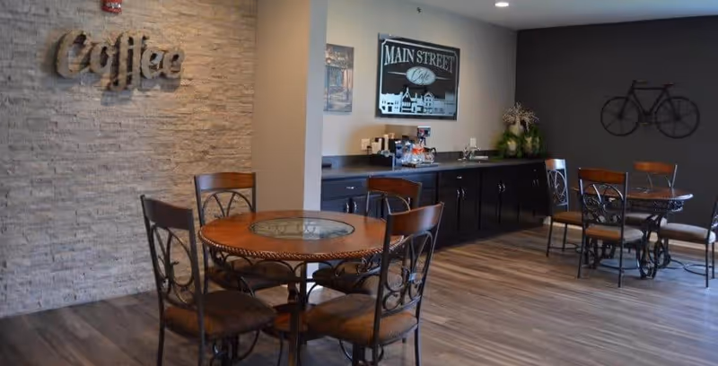 A cozy coffee area in a senior living community with round wooden tables and metal chairs. The wall on the left has a textured stone finish with the word 'Coffee' mounted on it. The back wall features a counter with coffee supplies and a framed sign that reads 'Main Street Cafe'. The right wall is dark with a decorative metal bicycle mounted on it. The floor is wood-style laminate.