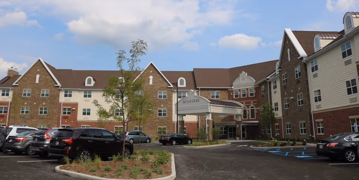 Exterior view of Belvedere Senior Housing building with a parking lot in front, several cars parked, and a covered entrance. The building has a mix of brick and siding with multiple windows and a clear blue sky with some clouds above.