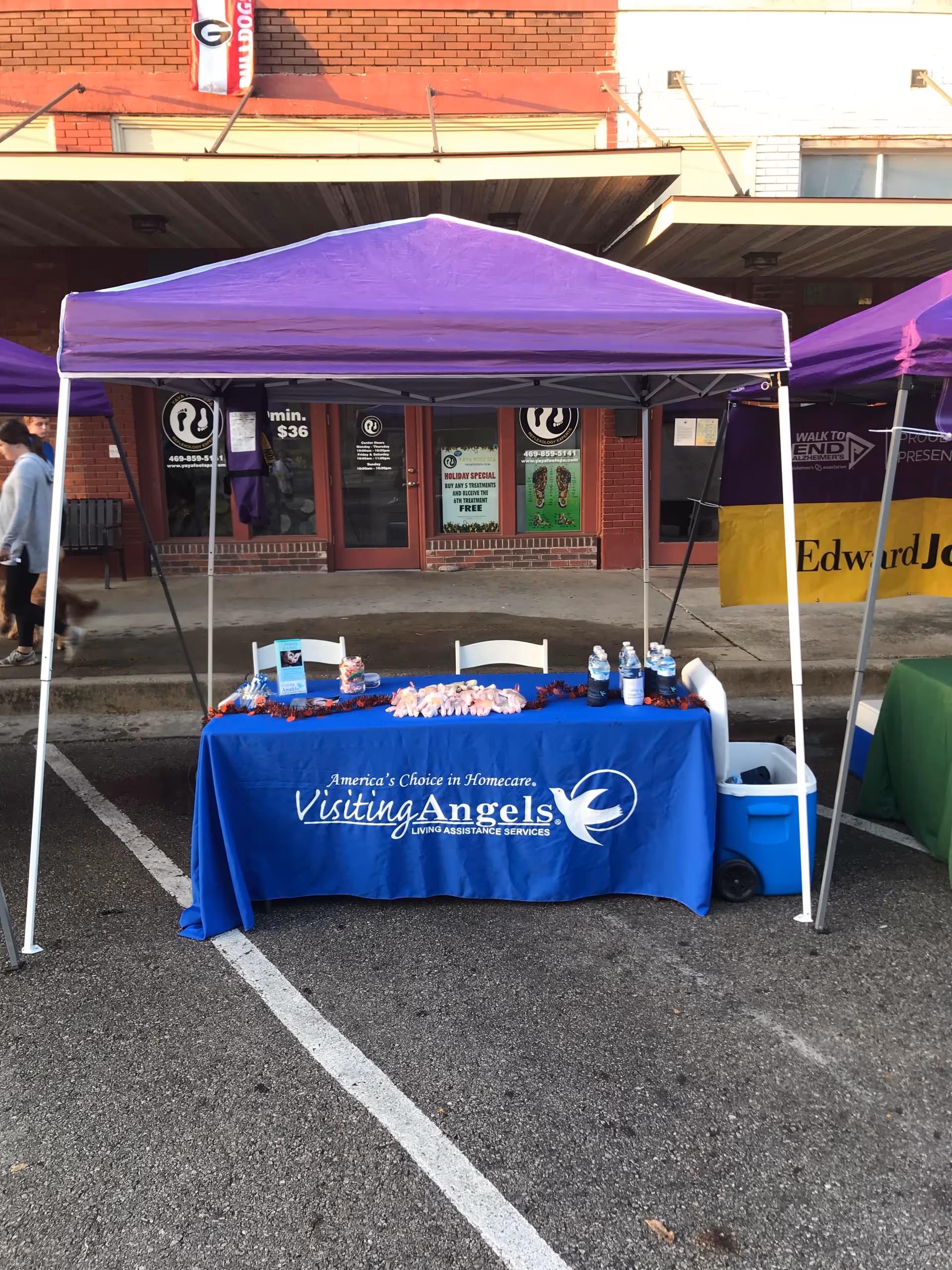 Outdoor promotional booth with purple canopy and blue tablecloth advertising Visiting Angels services.