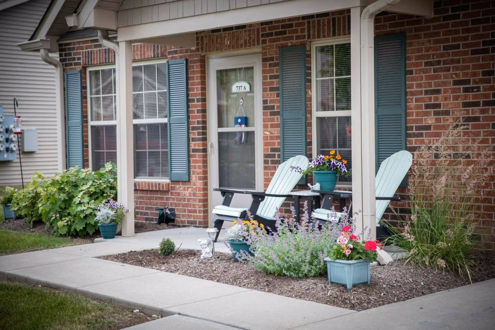 Front porch area of a brick building with two light blue Adirondack chairs and a small table between them. The porch is decorated with several potted plants and flowers. There are two windows with blue shutters on either side of a glass door labeled 737 A. A concrete walkway runs in front of the porch.