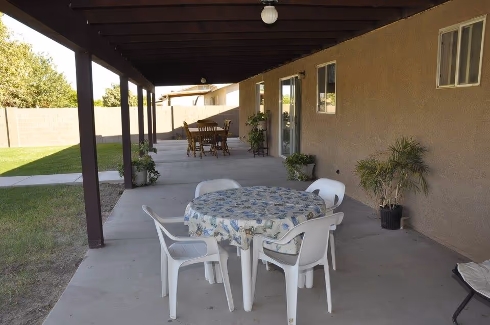 Covered outdoor patio area with a round table covered with a patterned tablecloth and four white plastic chairs. In the background, there is another wooden dining table with chairs. Several potted plants are placed along the wall of the building. The patio overlooks a grassy yard enclosed by a brick wall.