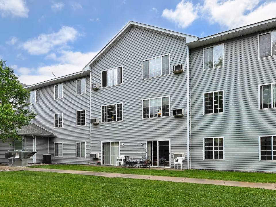 Exterior view of a three-story gray building with multiple windows and air conditioning units. There is a well-maintained green lawn in front with a concrete walkway and outdoor seating including chairs and a table. A tree is visible on the left side under a partly cloudy sky.