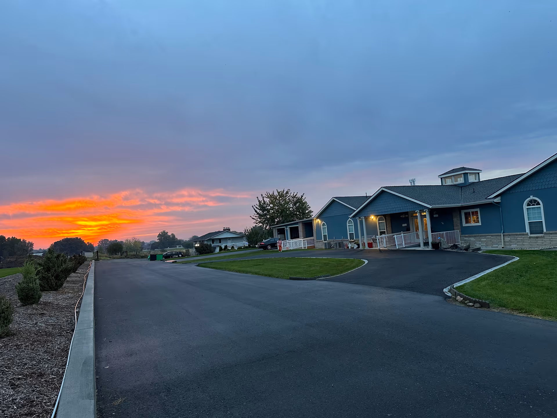 Sunset sky over blue adult family home with curved driveway and landscaped grounds.