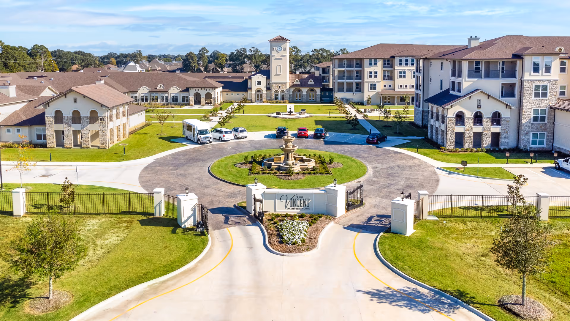 Aerial view of The Vincent Senior Living facility showing a gated entrance with a circular driveway and a central fountain. The buildings have a Mediterranean architectural style with beige walls, stone accents, and red-tiled roofs. There is a clock tower in the center background, surrounded by well-maintained lawns, trees, and parked vehicles.