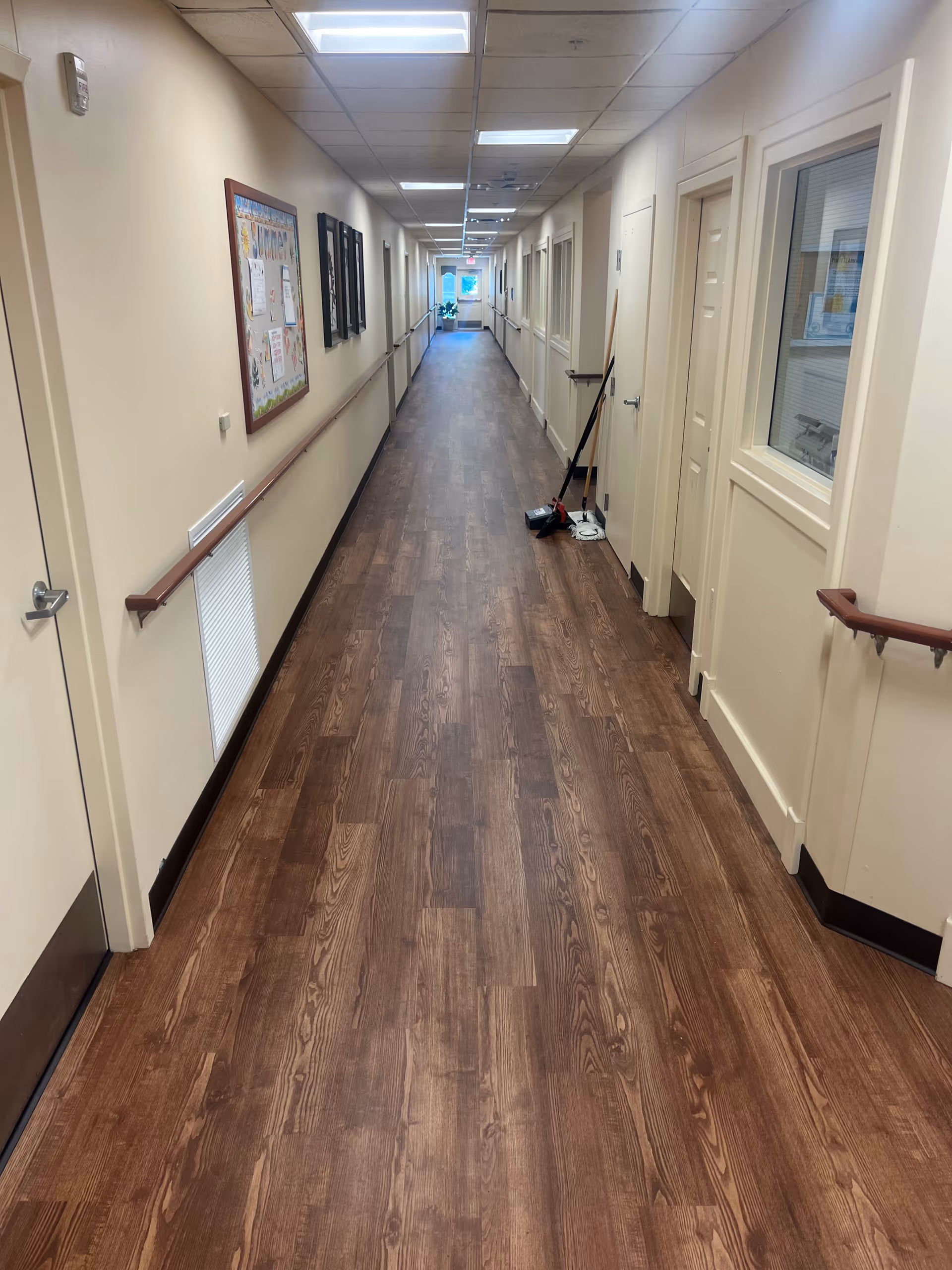 A long, clean hallway in a senior care facility with wood-patterned flooring, beige walls, handrails on both sides, several closed doors, a bulletin board on the left wall, and cleaning tools including a broom and mop leaning against the right wall.