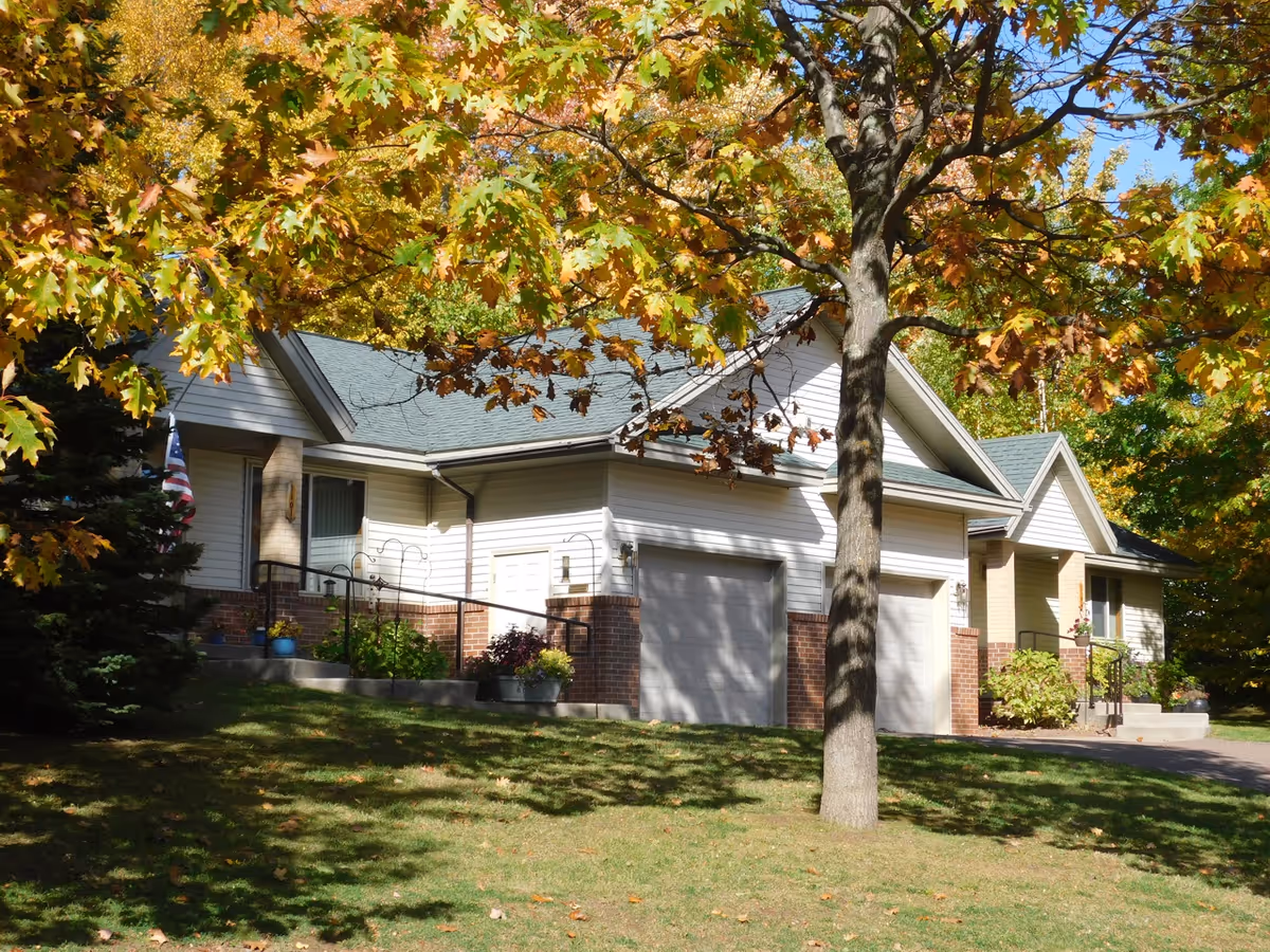 Front exterior of a one-story residential building with two garage doors, a large tree with autumn leaves, and a grassy lawn.