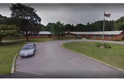 Brick senior living facility with a curved driveway, flagpole, and surrounding trees.