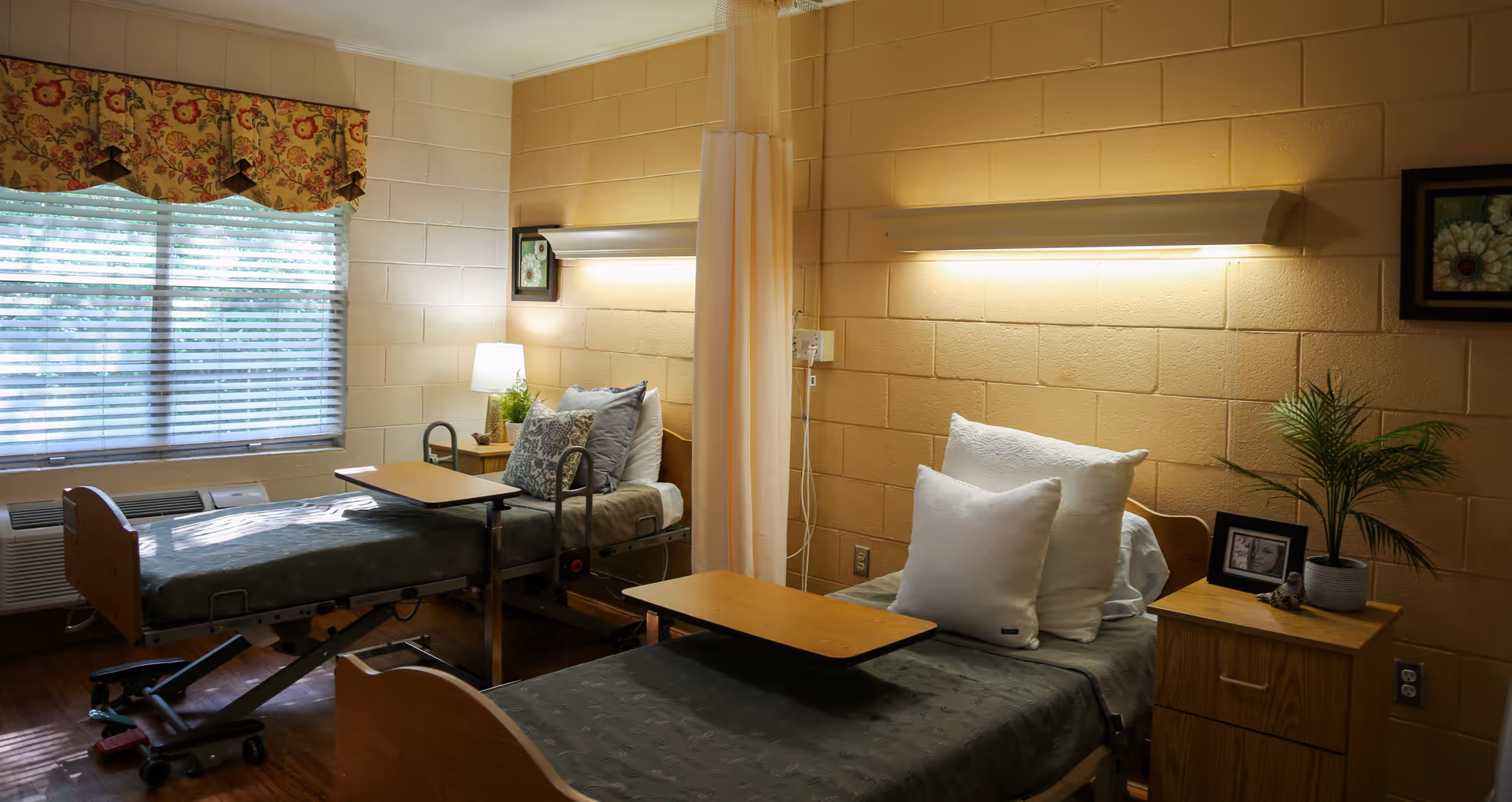 A shared patient room in a healthcare facility with two hospital beds separated by a privacy curtain. Each bed has pillows and a small overbed table. There is a wooden nightstand with a potted plant and framed photo between the beds. A window with blinds and a floral valance lets in natural light. The walls are painted beige with a textured finish, and there are wall-mounted lights above each bed.